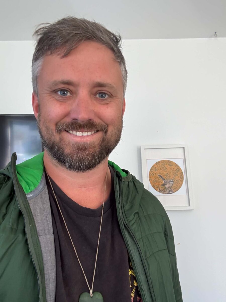 Tim MacDonald smiling, in a green jacket and a pounamu pendant.